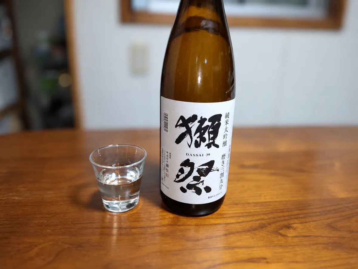 木目のテーブルに置かれた獺祭のボトルと、お酒が注がれた小さなグラス