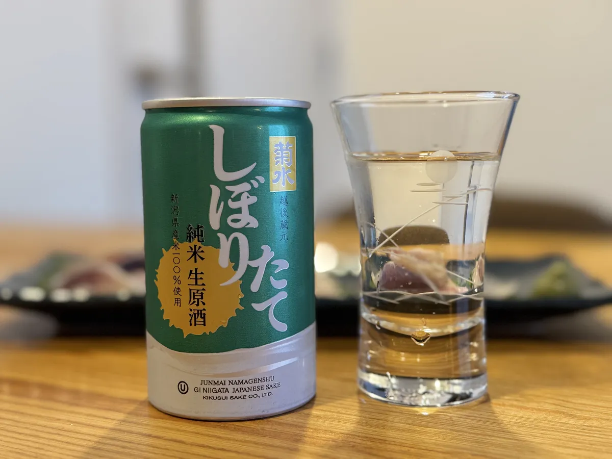 菊水 しぼりたて 純米生原酒のアルミ缶と、グラスに注がれたお酒、背景には刺身が並ぶ食卓