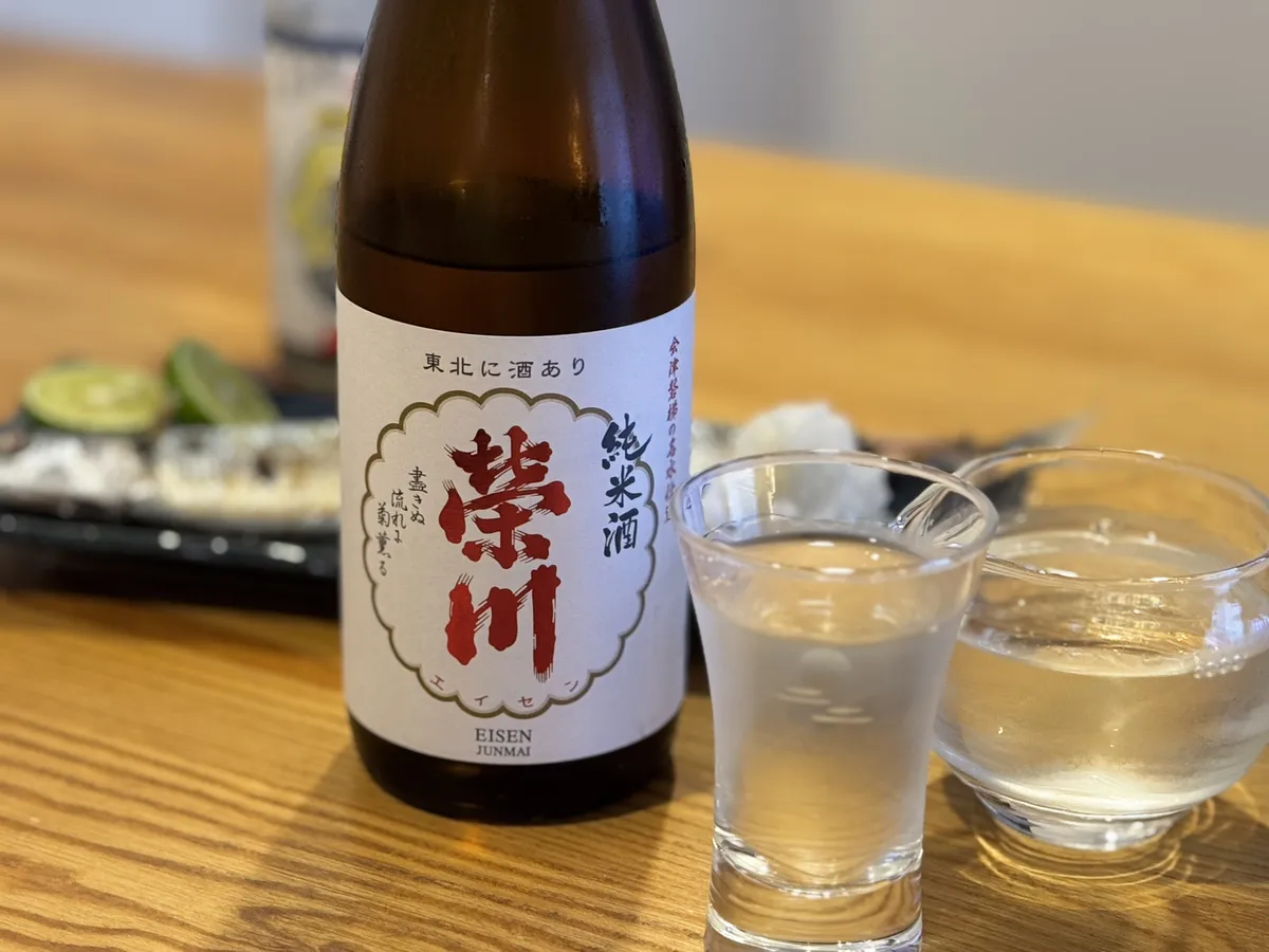木目のテーブルに置かれた榮川 純米酒のボトルと、秋刀魚の塩焼きが載った皿、お猪口に注がれたお酒