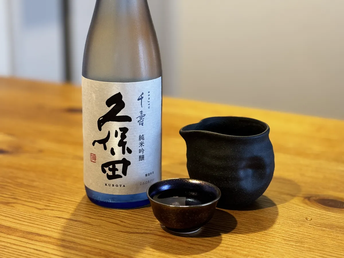 木目調のテーブルに置かれた久保田 千寿 純米吟醸のボトルと、黒い酒器に注がれたお酒