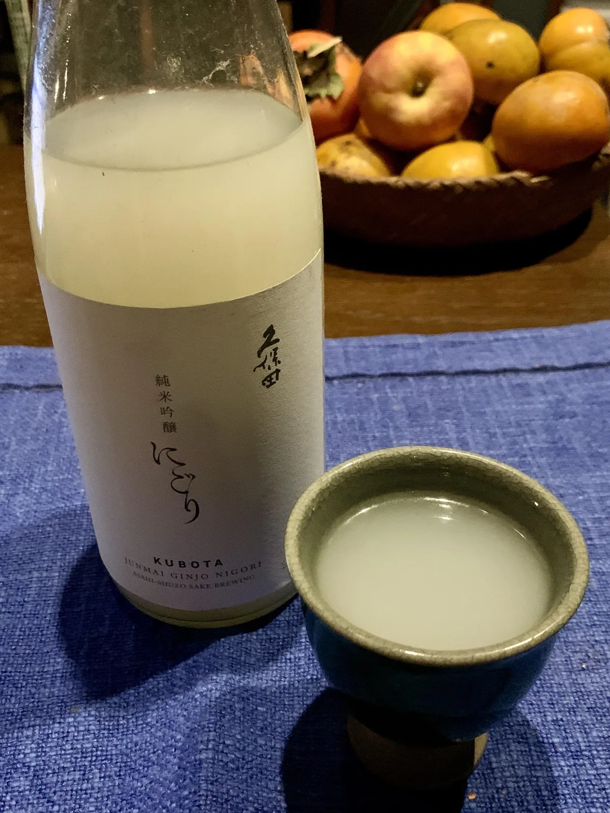 テーブルに置かれた久保田 純米吟醸 にごりのボトルと、注がれた酒器、背景に並ぶ果物
